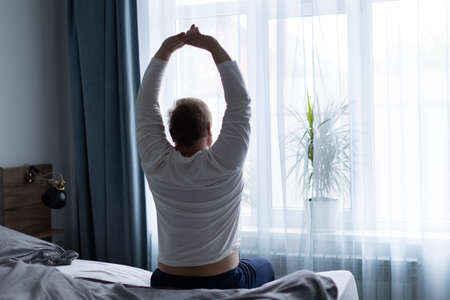 An adult man at the large window in the bedroom stretches in the morning after sleepingの写真素材