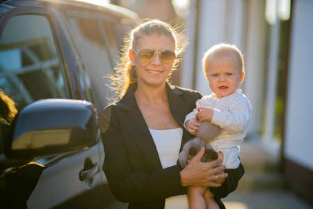 Beautiful business woman mother in a suit with a small child in her arms against the background of her house and a black car goes to workの写真素材