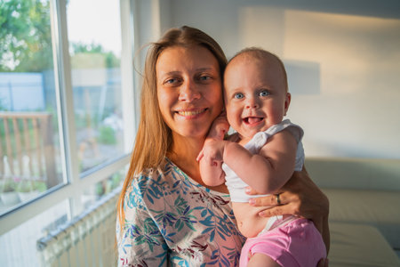 Portrait of a woman mother with a newborn baby in her arms at home next to a window in the room