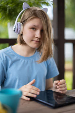 A teenage girl in a blue T-shirt in headphones with a tablet smartphone sits at a wooden table in a summer cafe and communicates on social networks and listens to musicの写真素材