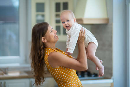 Beautiful young brunette woman in dress with baby in the kitchen at homeの写真素材