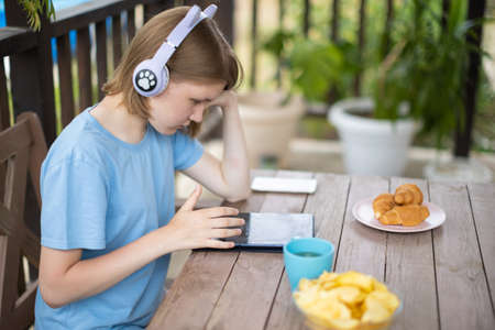 A teenage girl in a blue T-shirt in headphones with a tablet smartphone sits at a wooden table in a summer cafe and communicates on social networks and listens to musicの写真素材