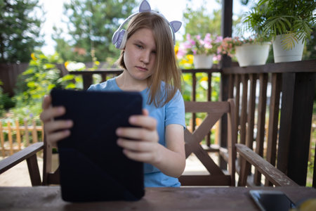 A teenage girl in a blue T-shirt in headphones with a tablet smartphone sits at a wooden table in a summer cafe and communicates on social networks and listens to musicの写真素材