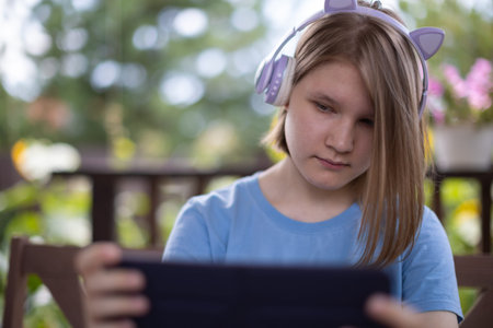 A teenage girl in a blue T-shirt in headphones with a tablet smartphone sits at a wooden table in a...
