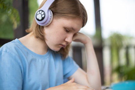A teenage girl in a blue T-shirt in headphones with a tablet smartphone sits at a wooden table in a summer cafe and communicates on social networks and listens to musicの写真素材