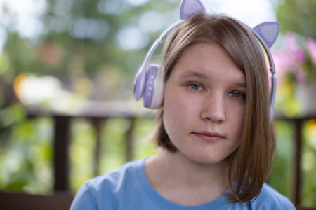 A teenage girl in a blue T-shirt in headphones with a tablet smartphone sits at a wooden table in a...