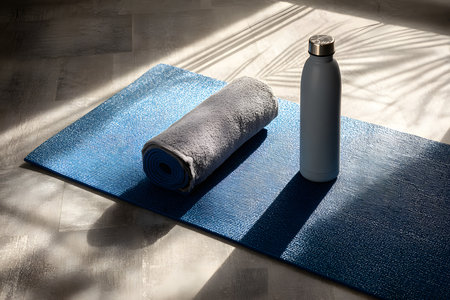 A blue yoga mat lies flat in a well-lit space. A rolled gray towel rests beside a white water bottle, creating an inviting atmosphere for morning exercise or relaxation.の素材
