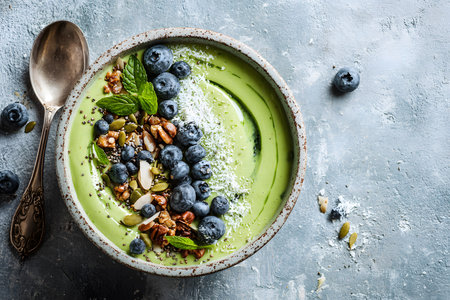 A vibrant green smoothie bowl is artfully topped with fresh blueberries, crunchy nuts, and mint leaves. The bowl rests on a textured gray surface, conveying a healthy breakfast option.の素材