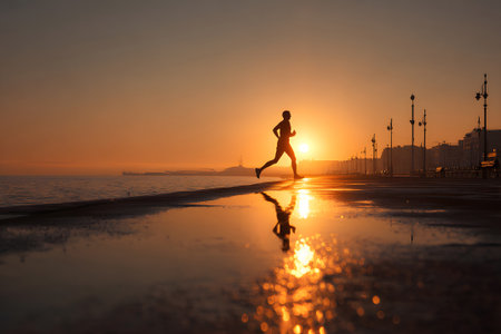 A person runs along a coastal path at sunrise, creating beautiful reflections on the wet pavement. The sky is filled with warm colors as day breaks over the horizon.の素材