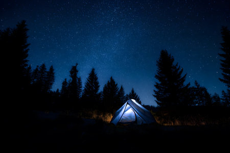 In a serene forest at night, a bright yellow tent emits a warm glow against a backdrop of twinkling stars and towering trees. Campers relax nearby, surrounded by nature's beauty.の素材