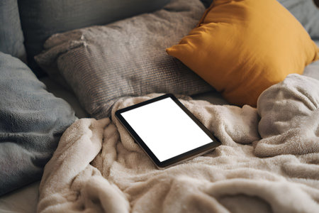 A modern bedroom scene shows a sleek tablet lying on a fluffy gray blanket amidst plush pillows, inviting relaxation and leisure time at home.の素材