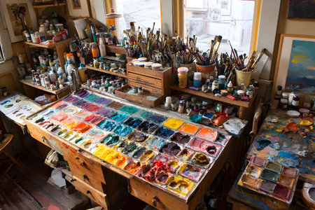 Bright sunlight filters through the window, illuminating a cluttered art table covered with paint jars, brushes, and a palette of vibrant colors, showing casing artistic inspiration.の素材