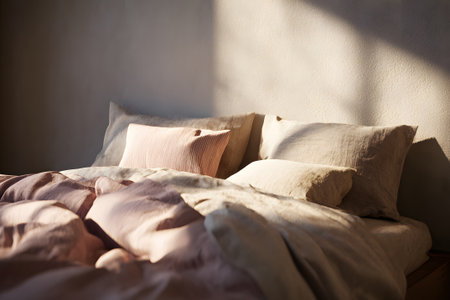 A well-made bed features light-colored pillows and a soft duvet. Sunlight filters through a window, adding warmth to the serene space. It invites relaxation and comfort.の素材