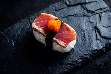 A single sushi roll sits on a dark stone surface, showing layers of rice, salmon, and sesame seeds. The simple presentation highlights the freshness and quality of the ingredients.の素材