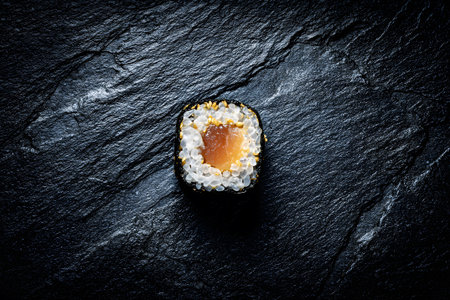 A single sushi roll sits on a dark stone surface, showing layers of rice, salmon, and sesame seeds. The simple presentation highlights the freshness and quality of the ingredients.の素材