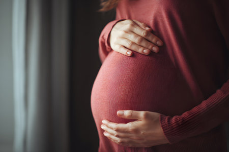 Pregnant woman gently cradles her baby bump while standing by a window in soft natural lightの素材