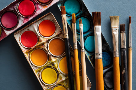Vivid watercolor paints in a palette sit beside various brushes on a rustic wooden surface. The scene captures the essence of a creative art session taking place in the afternoon.の素材