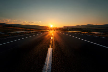 A long, straight road leads into the distance as the sun sets behind the mountains. The golden light bathes the landscape and highlights the pavement.の素材