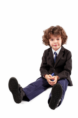 Curly boy in a jacket and tie sitting on the floor with legs stretched and looking at the camera. White background.の写真素材