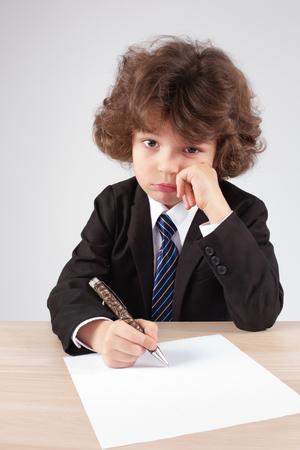 Little curly-haired boss is sadly looking at the camera with a blank sheet of paper and a pen. Gray background. Close-up.の写真素材