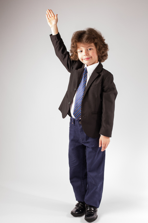 Funny curly boy raised his hand in a business suit, smiling and looking at the camera. Full length. Gray background.の写真素材