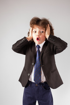 Funny curly boy in a business suit clutched his hands behind his head and his mouth open in amazement looking at the camera. Grey background.の写真素材
