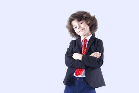 Curly cute boy in a business suit with his arms folded, his head bowed looking at the camera. Gray background.の写真素材