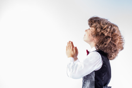 Little curly boy with her hands folded in prayer, looking upwards. Sideways to the camera. White background.の写真素材