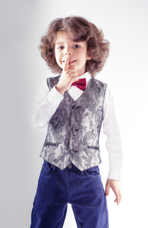 Funny curly-haired boy in waistcoat showing gesture "Do not make a noise." Gray background.の写真素材
