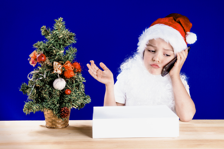 Young Santa Claus sitting at the table, sticking out his lip and spreading hands on the phone. White box with gifts and a Christmas tree on a table. Blue background. Chromakey. Close-up.の写真素材
