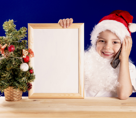 Young Santa Claus sitting at a table, laughing and talking on the phone. Keep empty frame with a white background. He looks into the camera. Christmas tree on the table. Blue background. Close-upの写真素材