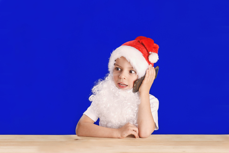 Young Santa Claus sitting at a wooden table and talking on the phone. Surprised looking at the Christmas tree. Blue background. ?hromakey. Close-up.の写真素材