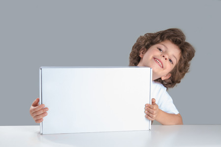 Cute curly little boy sitting at a table holding a white box. He looks into the camera. Gray background. Close-up.の写真素材