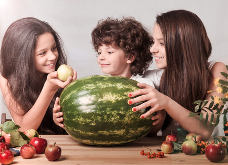 Three children playing with fruits sitting at the table in front of a huge watermelon. Gray background. Close-up.の写真素材