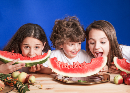 Little curly boy and two girls sitting at a wooden table and eat large ripe watermelon. Blue background. Close-up.の写真素材