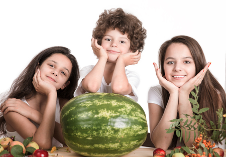 Two girls and a boy with his head on his hands, sitting at the table in front of a large watermelon whole. Looking at the camera. White background. Close-up.の写真素材