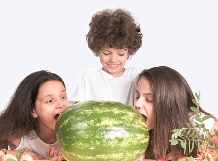 Two sisters bite a huge watermelon. Brother looking down at them. White background. Close-up.の写真素材