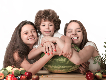 Three happy children hugging a huge watermelon, sitting at the table. Smiling, looking into the camera. White background. Close-up.の写真素材
