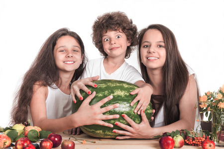 Three happy children holding hands on a huge watermelon, sitting at the table. Smiling, looking into the camera. White background. Close-up.の写真素材