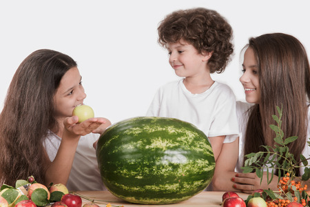 Brother and two sisters looking at each other sitting at the table. On the table is a huge whole watermelon and fruit. White background. Close-up.の写真素材
