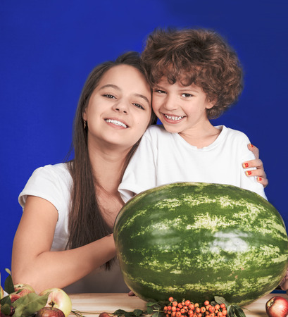 Sister hugging her brother sitting at the table in front of a large watermelon whole. Looking at the camera. Blue background. Close-up.の写真素材