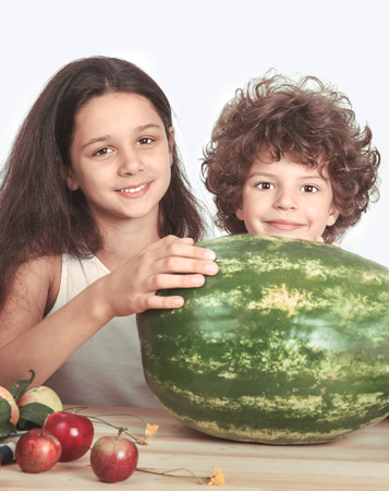 Curly boy and a girl sitting at a table in front of ripe watermelon. Looking at the camera. Grey background. Close-up.の写真素材