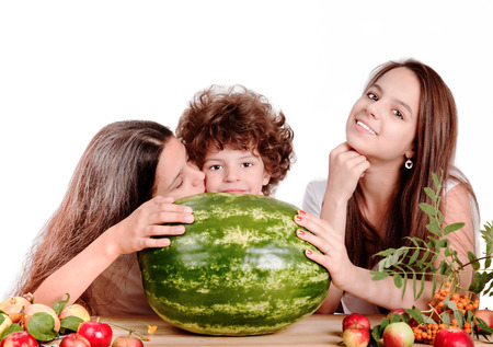 Brother and two sisters are sitting at the table in front of ripe watermelon. A girl kisses a boy on the cheek. White background. Close-up.の写真素材