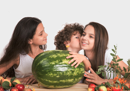 Three children sitting at the table. The boy put his hand on a large watermelon and whispers in his ear a girl. Gray background. Close-up.の写真素材
