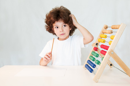 Cute curly-haired boy with a pencil in his hand thoughtfully scratching his head and looking at the camera. Close-up. Gray background.の写真素材