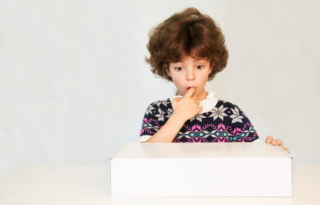 Thoughtful cute young boy in a sweater with his finger in his mouth, stares at the white box. Gray background.の写真素材