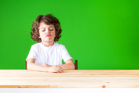 Curly boy sticking out his lips in disbelief looks at the table. Close-up. Green background.の写真素材