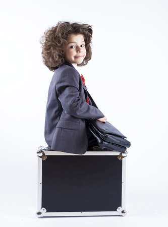 Little curly-haired boy dressed in a business suit sitting on the black suitcase looking at the camera. Isolated on a gray background.の写真素材