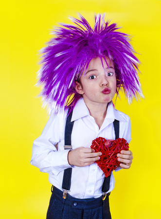 Beautiful baby with a lilac party wig holding a red heart on a yellow backgroundの写真素材