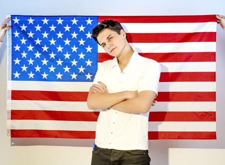 Young confident man in white shirt with arms crossed, standing against US flag background who hold two handsの写真素材